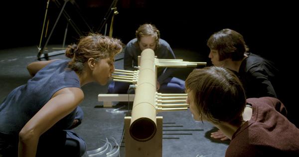 A group of performers blow into a large flute like instrument - the piece is called Coil of Days Scene 2 Communal Breath by Rita Evans