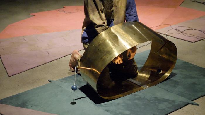 An artist kneels underneath a wide strip of copper bent into an oval shape on the ground - the photo captures a moment in Rita Evans' Tuning in a Vacuum piece
